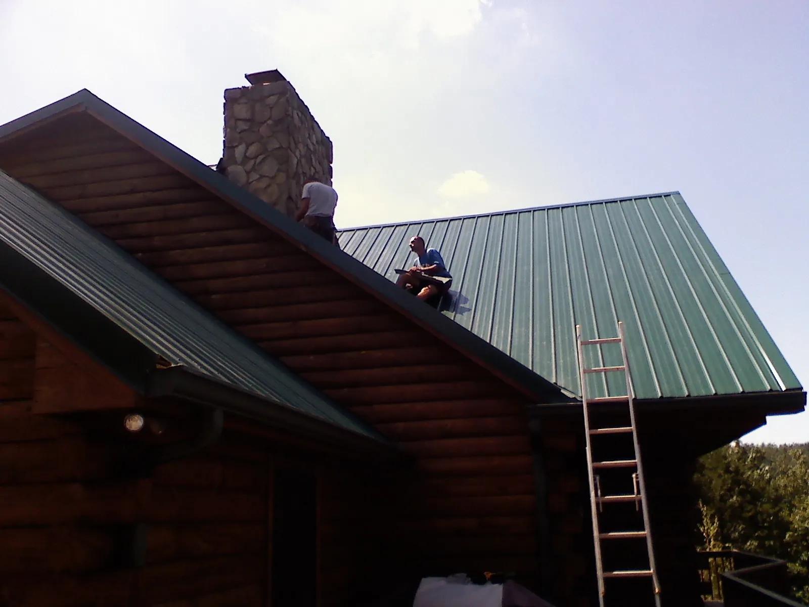 Roofers installing green metal panels beside a stone chimney on a log home in Bridgeton